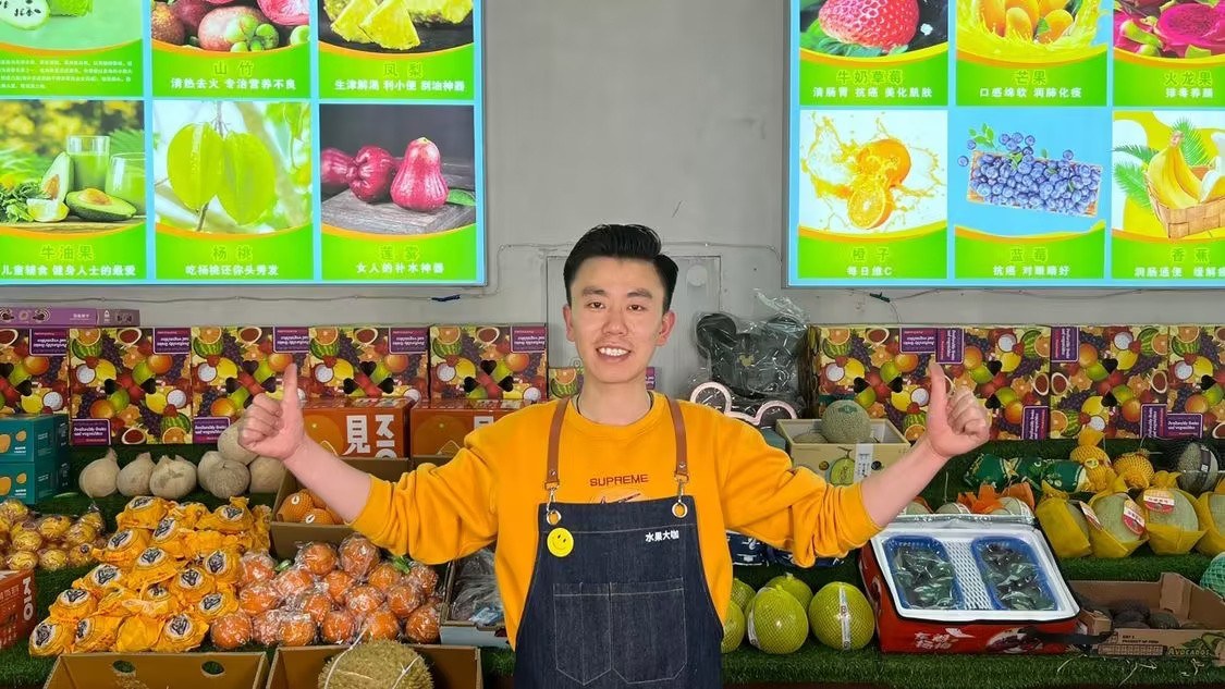 王岐站在自己的精品水果店里，身后是堆满的水果和水果灯箱广告
