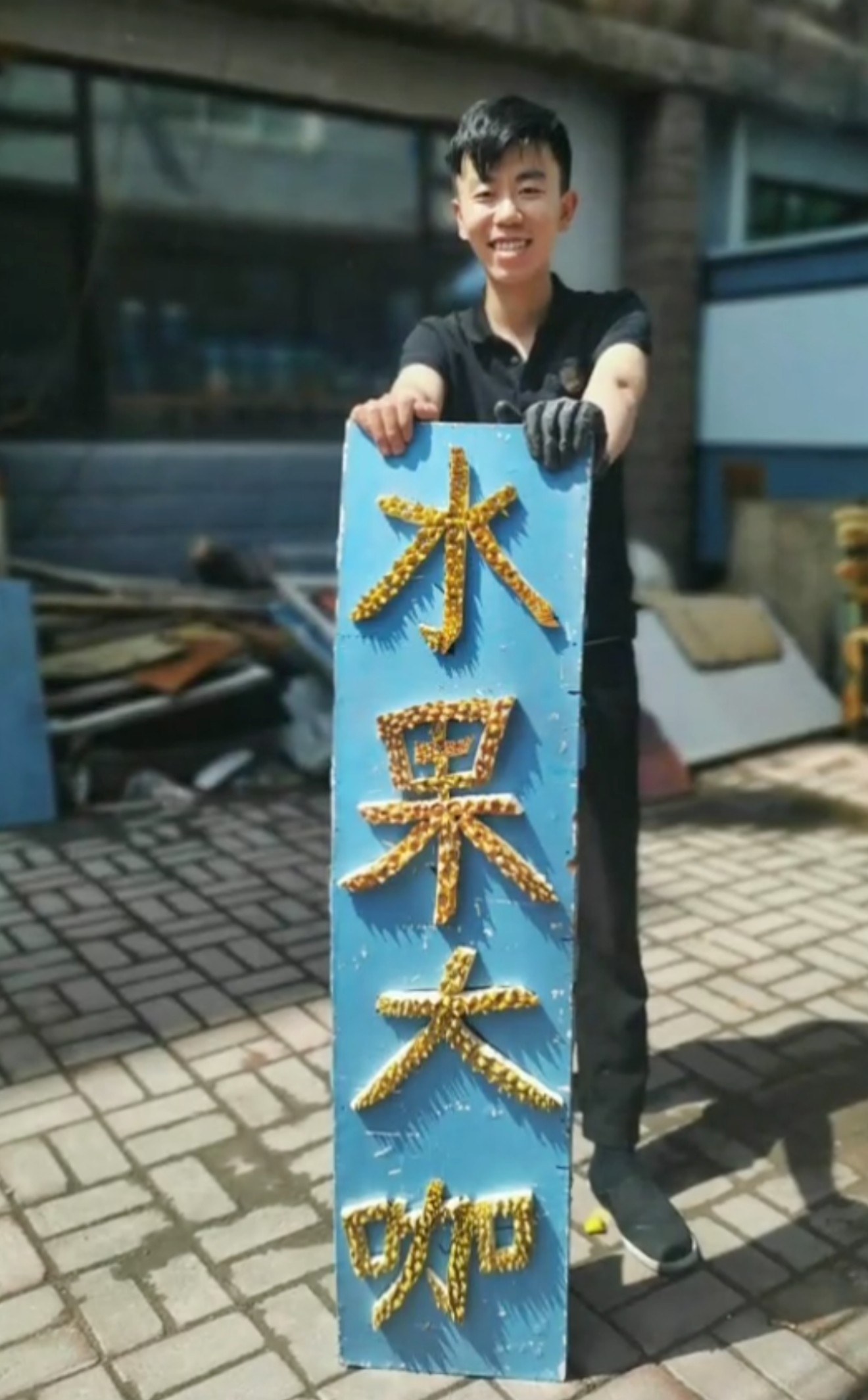 王岐双手扶着一块蓝色招牌，招牌上用榴莲刺壳拼出“水果大咖”四个字