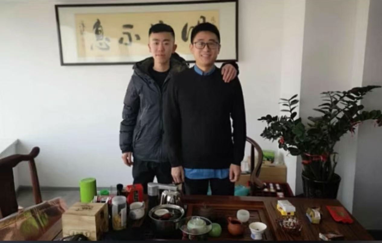 十年前大飞和王岐的第一张合影，两人站在一张摆着茶具的茶桌后，身后墙上挂着书法